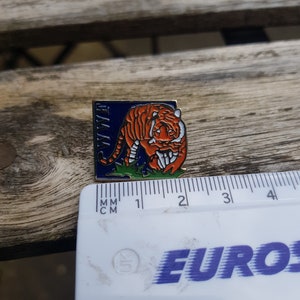 May include: A blue and green enamel pin featuring a tiger with orange and white stripes. The pin is approximately 1.5 inches tall and says "WWF" on the side.