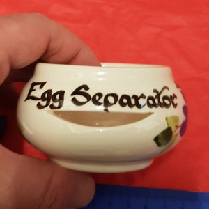 Puede incluir: Separador de huevos de cerámica blanca con un diseño floral y las palabras "Egg Separator" escritas en letra negra.