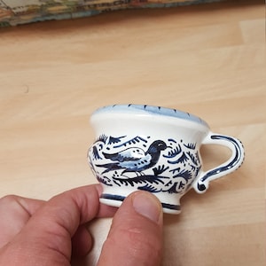 Pu&ograve; includere: Una piccola tazza in ceramica bianca con un motivo floreale blu e bianco e un uccello. La tazza ha un manico e una base arrotondata.