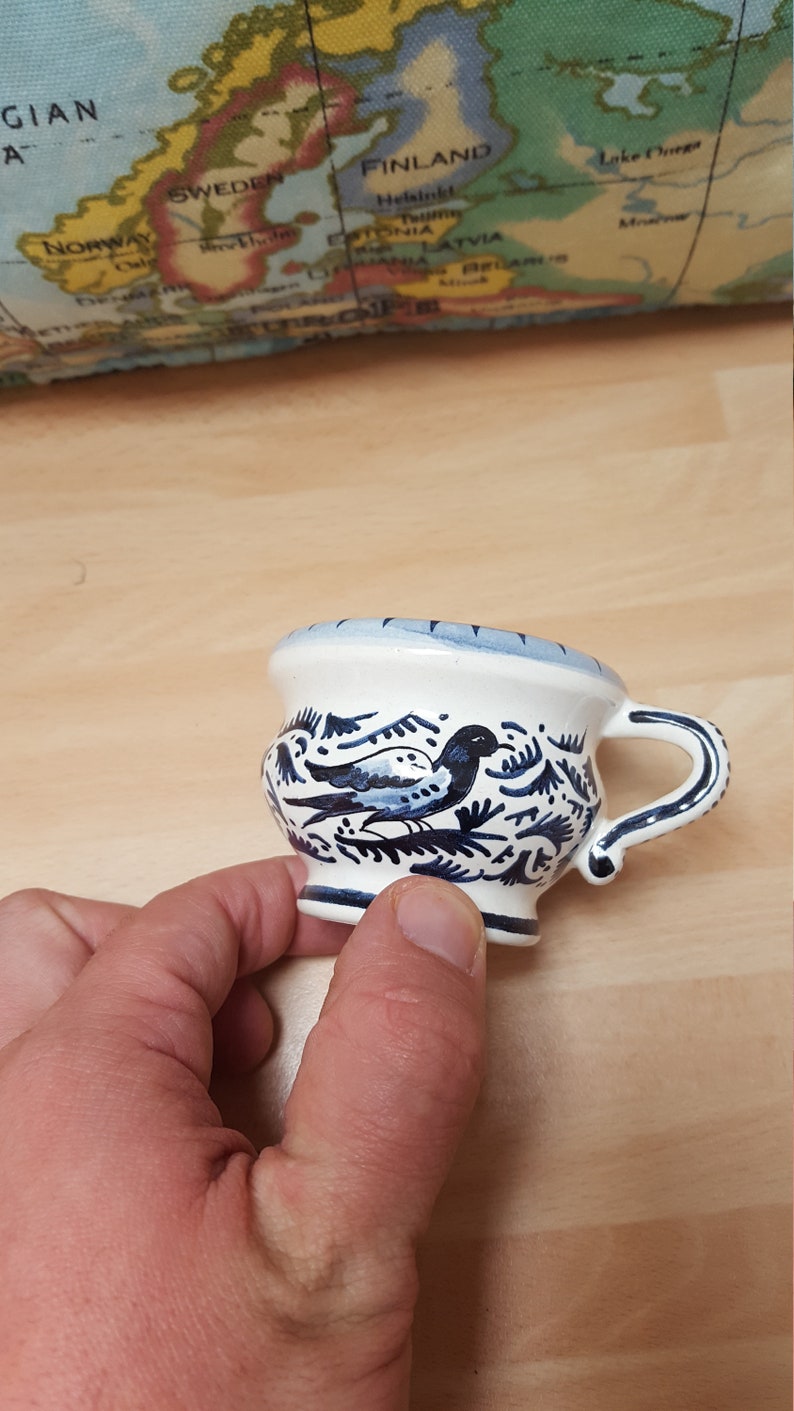 Pu&ograve; includere: Una piccola tazza in ceramica bianca con un motivo floreale blu e bianco e un uccello. La tazza ha un manico e una base arrotondata.
