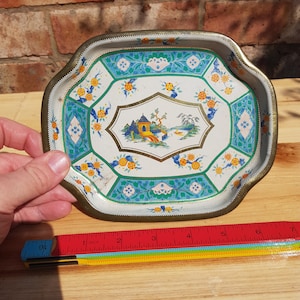 Puede incluir: Una bandeja de metal blanco con un borde festoneado y un diseño floral colorido. La bandeja presenta una imagen central de una pagoda y un puente sobre un río con un barco en el agua. La bandeja tiene un diámetro de aproximadamente 15 cm.