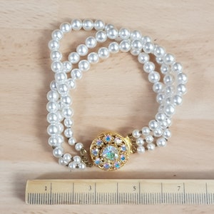 Puede incluir: Un brazalete de perlas blancas con un cierre dorado y pedrería de cristal de colores. El brazalete mide aproximadamente 12,7 cm de largo.