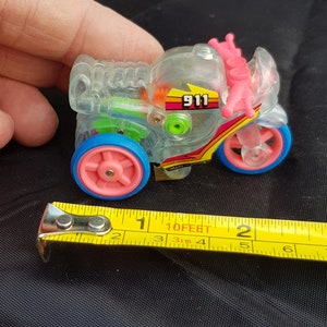 Puede incluir: Una motocicleta de juguete de plástico transparente con detalles en rosa y azul. La motocicleta tiene un diseño de rayas amarillas y rojas y el número 911 impreso en el lateral. La motocicleta mide aproximadamente 5 cm de largo.