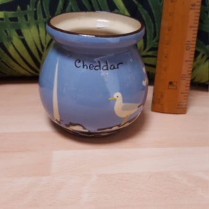May include: A small blue ceramic jar with a white rim and a seagull design. The jar has the word "Cheddar" written on it.