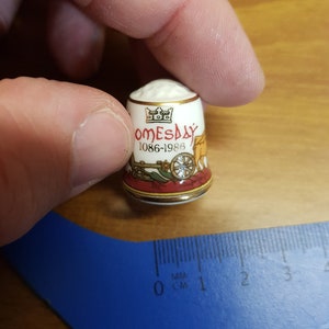 Può includere: Ditale in ceramica bianca con bordatura dorata e design rosso, giallo e nero. Il ditale presenta una corona e il testo "Domesday 1086-1986".