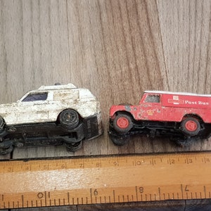 May include: Two toy vehicles, a white van and a red post bus, are side-by-side on a wooden surface. The vehicles are both slightly weathered and have a vintage look.