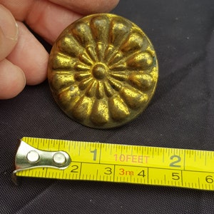 May include: A gold-colored metal button with a raised floral design. The button is approximately 1 inch in diameter.
