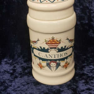 Op de afbeelding: Witte keramische pot met een blauw en gouden deksel. De pot heeft een blauw en gouden etiket met de tekst "C. ANTHOS" en een cherubijn met vleugels. De pot is versierd met bloemen en vogels.