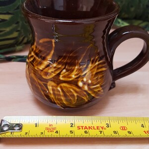 May include: A brown ceramic mug with a brown and yellow floral design. The mug has a handle and is approximately 10 cm tall.