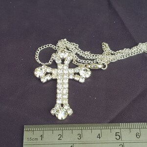 May include: A silver chain necklace with a large rhinestone cross pendant. The cross is approximately 5 cm tall and is covered in clear rhinestones. The chain is about 45 cm long.