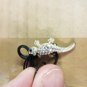 May include: A silver-toned lizard pendant, encrusted with clear crystals, is attached to a black cord. The lizard has detailed features and is positioned on a light brown surface. The pendant is a jewelry item.