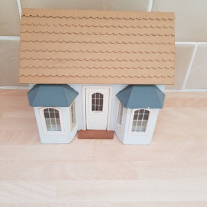 May include: A white dollhouse with a brown shingled roof, blue trim, and three windows. The dollhouse has a front door and a small porch.