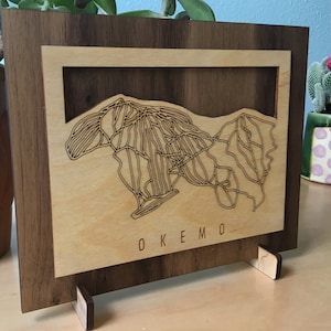 May include: A wooden wall art piece with a laser-cut design of a mountain range. The art piece is framed in a darker wood and has a stand on the back. The text "OKEMO" is engraved below the mountain design.