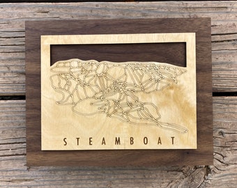 May include: Wooden wall art featuring a detailed map of Steamboat ski resort. The map is laser-cut into a light-colored wood, set within a dark wood frame. The word "STEAMBOAT" is engraved at the bottom of the map.