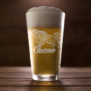 May include: A tall glass filled with beer and a thick foamy head. The glass is etched with a mountain design and the word "Okemo". The glass is covered in condensation, sitting on a wooden surface.