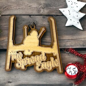 May include: A wooden Christmas ornament shaped like a skier in the spread eagle position. The ornament is brown with a light brown skier silhouette and the text "The Spread Eagle" in a decorative font.