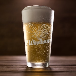 Könnte beinhalten: Ein hohes Glas gefüllt mit Bier und einer dicken weißen Schaumkrone. Auf dem Glas sind das Wort "Windham" und eine Berg-Grafik eingraviert. Das Glas ist mit Kondenswasser bedeckt und steht auf einer Holzoberfläche.