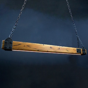 May include: A rustic wooden pendant light fixture with black metal chains and brackets. The wood has a natural grain pattern and is finished in a light brown color. The light fixture is suspended from the ceiling by two black chains.