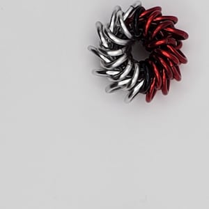 Op de afbeelding: Twee zilveren en rode metalen ringen, elk met een zwart midden, worden getoond tegen een witte achtergrond. De ringen zijn gemaakt van kleine, met elkaar verbonden metalen lussen.