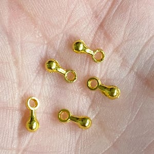 May include: A close-up of six gold-colored metal bead caps. The bead caps are in the shape of a teardrop with a small loop at the top. The bead caps are on a person's hand.