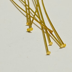 May include: A group of ten gold-colored head pins with rounded ends. The pins are arranged in a fan shape on a white surface.