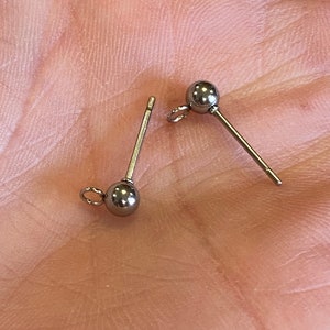 May include: Two silver earring posts with small round balls at the end. The posts are on a hand.