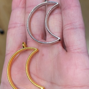 May include: Two crescent moon shaped pendant bezels, one in a polished silver tone and one in a gold tone. Each open frame features a small circular loop at the top for attachment, designed for jewelry making or crafting projects.