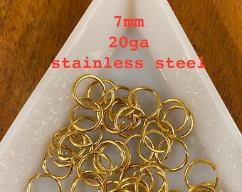 Puede incluir: Una colección de anillas de salto doradas de acero inoxidable, cada una de 7 mm de diámetro y 20ga de grosor. Los anillos metálicos se muestran en una bandeja triangular blanca, dejando ver su forma circular y diseño abierto.