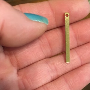 May include: A small, rectangular, gold-colored metal bar with a hole at the top. The bar is held in a person's hand with a turquoise painted fingernail visible in the background. The bar appears to be a jewelry component or finding.