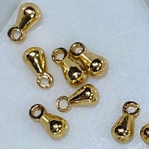 May include: A group of ten gold-colored teardrop-shaped charms with a loop at the top. The charms are arranged on a white surface.