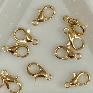 May include: A close-up of nine gold-toned lobster clasps, a type of jewelry finding, displayed on a white triangular surface.