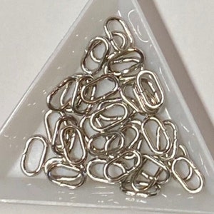 May include: A white triangular tray filled with silver jump rings. The jump rings are small and oval shaped.