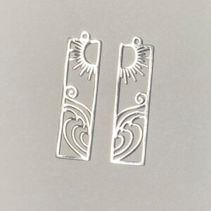 May include: Two silver-colored rectangular earrings with a sun and wave design. The top of each earring features a sun with rays, and the bottom has a stylized wave pattern. The earrings are lightweight and have a small loop at the top for hanging.