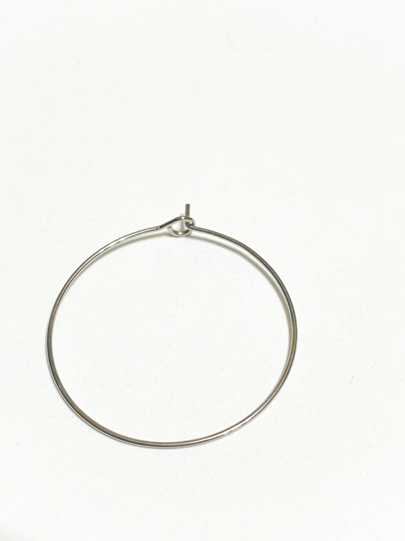 Op de afbeelding: Een dunne, cirkelvormige oorring van zilverkleurig metaal. De draad vormt een volledige cirkel met een kleine lus aan de bovenkant waar de uiteinden samenkomen, voorzien van een recht staafje dat iets omhoog steekt uit het sluitmechanisme.