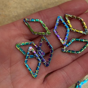May include: Iridescent metal diamond-shaped charms. The charms have a geometric design with a color gradient of green, blue, and purple. The charms are held in a hand.