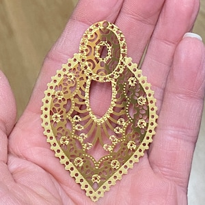 May include: A gold-colored filigree earring with an intricate openwork design. The earring features a teardrop shape with a decorative oval top and a scalloped edge. The delicate pattern includes floral and geometric elements.