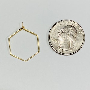 Puede incluir: Un pendiente dorado, geom&eacute;trico, en forma de hex&aacute;gono, junto a una moneda de veinticinco centavos estadounidense. El pendiente est&aacute; hecho de alambre fino y tiene un dise&ntilde;o simple y minimalista. La moneda es plateada y muestra el perfil de una persona.