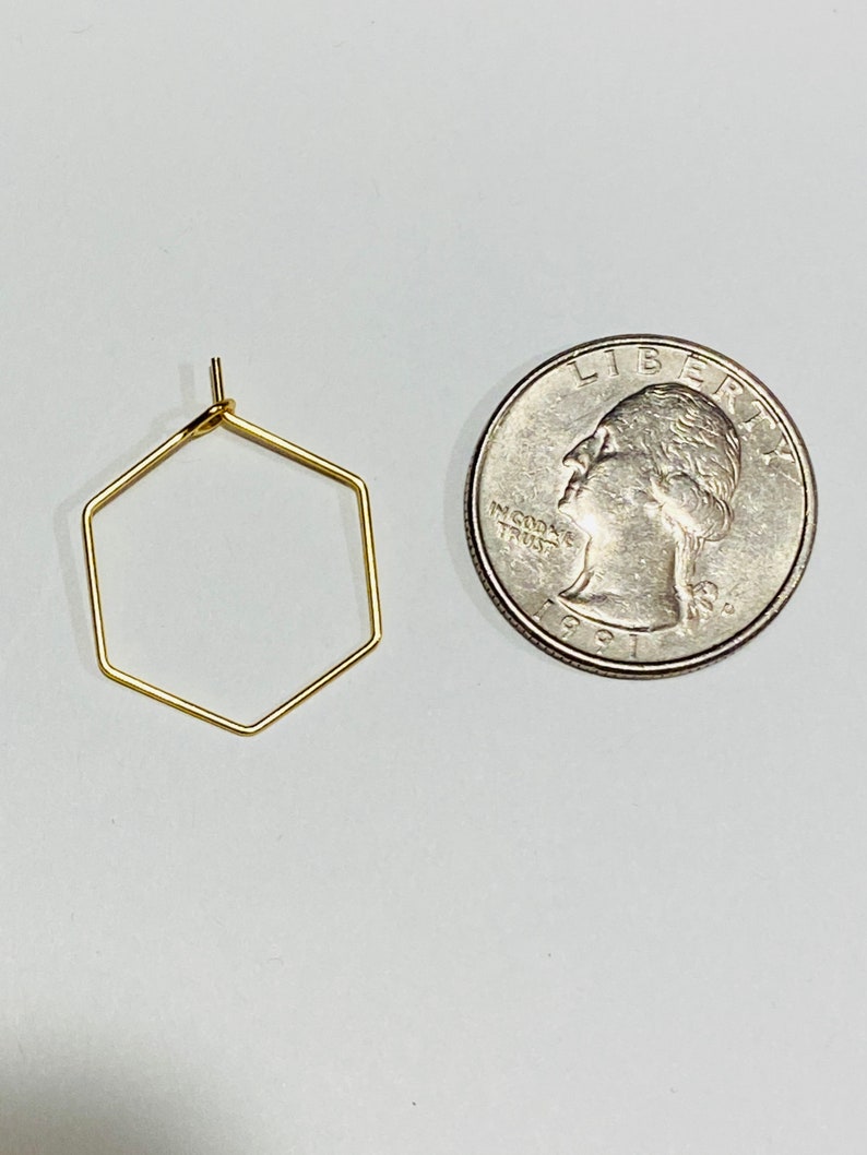 Puede incluir: Un pendiente dorado, geom&eacute;trico, en forma de hex&aacute;gono, junto a una moneda de veinticinco centavos estadounidense. El pendiente est&aacute; hecho de alambre fino y tiene un dise&ntilde;o simple y minimalista. La moneda es plateada y muestra el perfil de una persona.