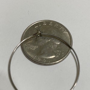 Op de afbeelding: Een dunne, cirkelvormige zilverkleurige metalen oorring geplaatst over een Amerikaanse kwartdollar uit 1991. De ring is voorzien van een eenvoudig draadsluitingsmechanisme aan de bovenkant en de munt dient als visuele schaal voor de maat.