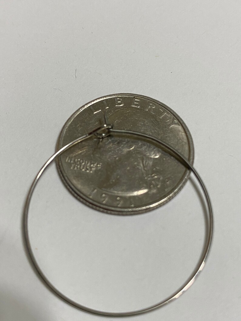 Op de afbeelding: Een dunne, cirkelvormige zilverkleurige metalen oorring geplaatst over een Amerikaanse kwartdollar uit 1991. De ring is voorzien van een eenvoudig draadsluitingsmechanisme aan de bovenkant en de munt dient als visuele schaal voor de maat.