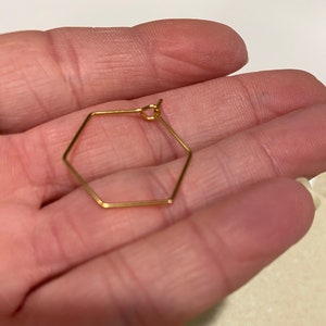 Puede incluir: Un solo pendiente hexagonal de color dorado. El pendiente tiene una forma geom&eacute;trica con un dise&ntilde;o delicado y minimalista. El pendiente est&aacute; sostenido en la mano de una persona.