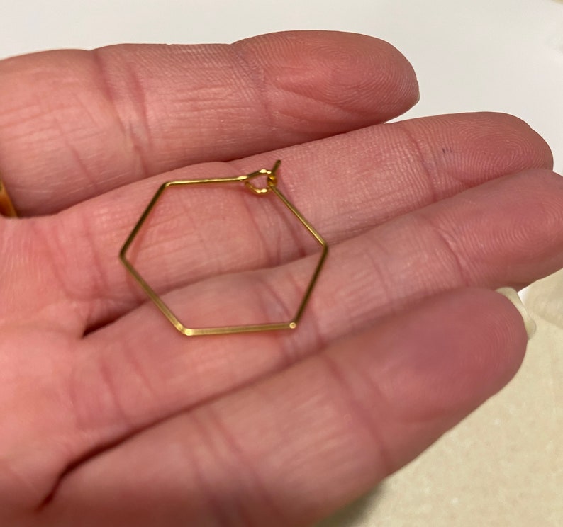 Puede incluir: Un solo pendiente hexagonal de color dorado. El pendiente tiene una forma geom&eacute;trica con un dise&ntilde;o delicado y minimalista. El pendiente est&aacute; sostenido en la mano de una persona.