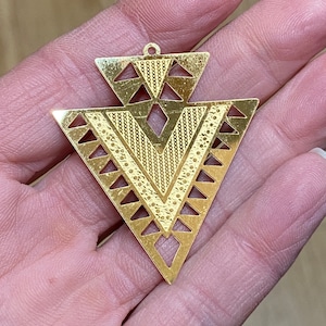 May include: Gold-toned metal pendant with a geometric design featuring a large triangle with a smaller triangle at the top. The pendant has a cutout design and a textured surface.