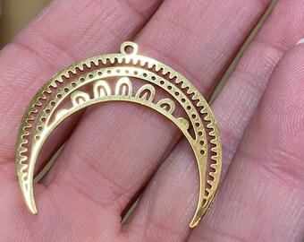 May include: A gold-colored metal crescent moon pendant featuring intricate cut-out details, including a serrated outer edge, a row of small circular perforations, and a series of arched shapes along the inner curve with a small loop at the top.