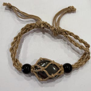 May include: A handmade bracelet featuring a central dark, oval-shaped stone encased in a woven net. Two black, round beads flank the center stone. The bracelet is crafted from a natural-colored, braided cord, with adjustable ties.