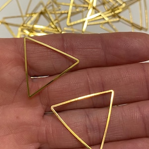 May include: Two gold-colored metal triangle shapes, one larger than the other, are shown on a hand. The triangles are open and have a smooth, shiny finish.