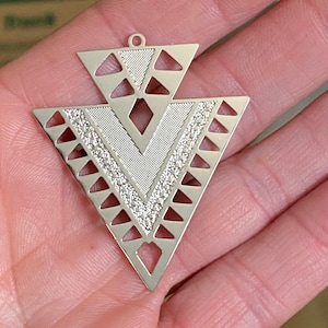 May include: Silver pendant with a geometric design featuring a large triangle with a smaller triangle inside. The pendant has a textured surface and a cut-out design.