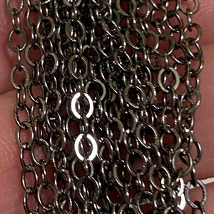May include: Close-up of a dark gray chain. The chain is made of small, round links, creating a textured surface. The links are closely connected, forming multiple strands. The chain has a metallic sheen, reflecting light.