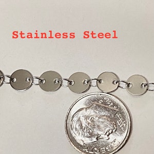 May include: A close-up of a silver chain made of small, round, connected links. The chain is next to a 10 pence coin for scale. The text "Stainless Steel" is visible at the top of the image.