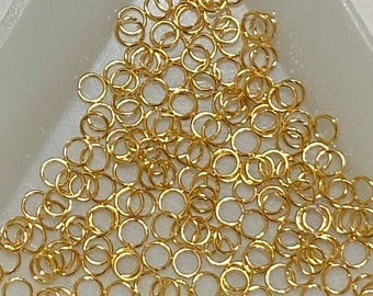 Puede incluir: Una colección de pequeñas anillas de metal de color dorado dispuestas en formación triangular. Cada anilla individual presenta una estructura de alambre fino con una abertura visible, comúnmente utilizada como conector en joyería y manualidades.